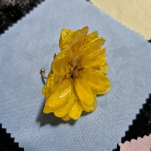 Anillo ajustable de acero con flor real de dalia amarilla encapsulada en resina brillante sobre un paño de microfibra celeste.