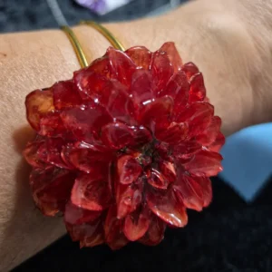 Brazalete rígido de acero inoxidable dorado con flor de dalia natural de color rojo anaranjado deshidratada y encapsulada en resina brillante sobre una muñeca.