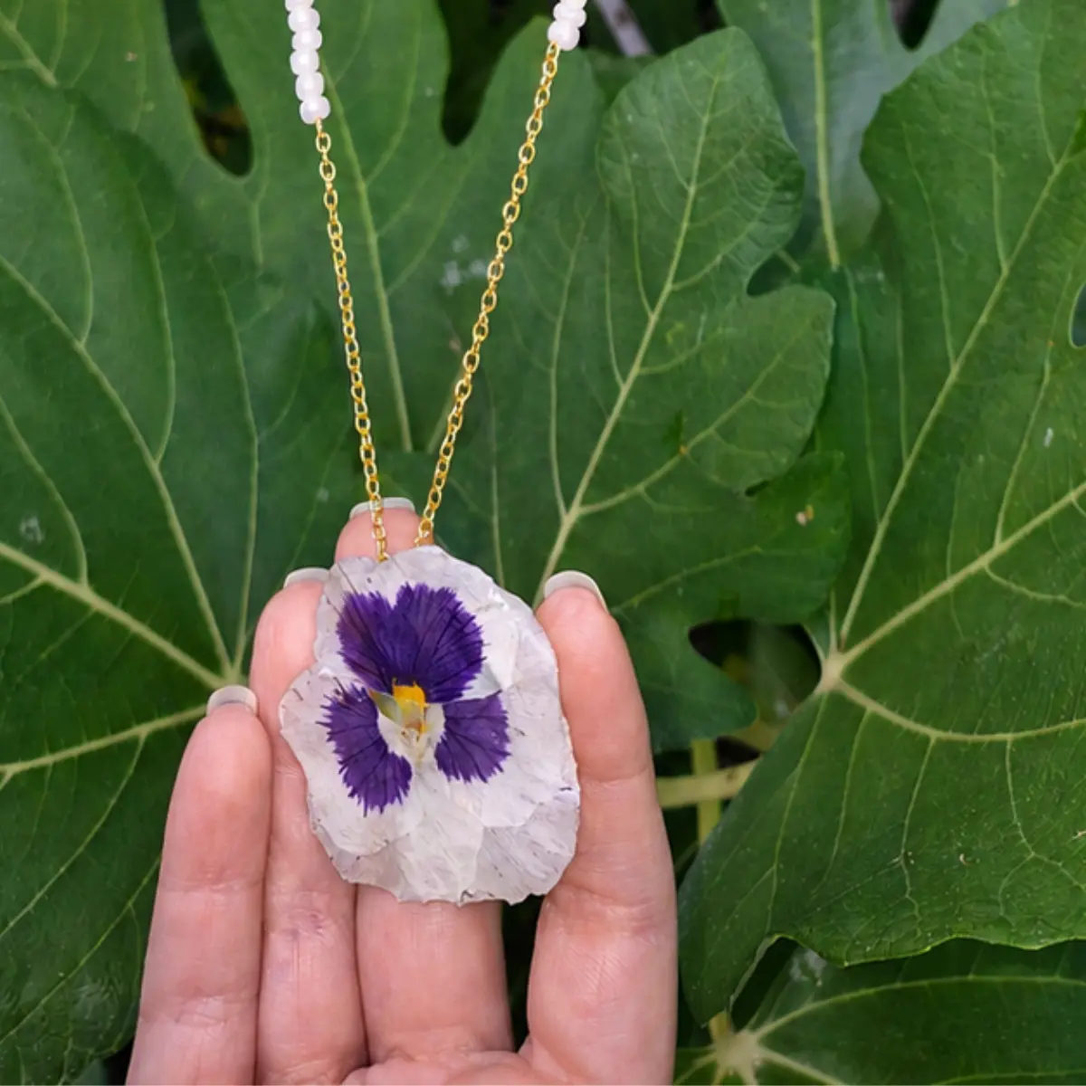 Collar de cadena dorada de acero inoxidable con colgante de flor real de pensamiento bicolor y cuentas de perlas irisadas expuesto ante un fondo de hojas verdes.