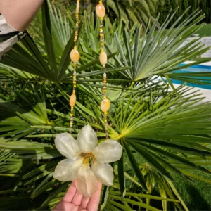 Manos sosteniendo en el aire un gran collar statement con un tulipán natural abierto encapsulado en resina transparente, combinado con una cadena de ágatas amarillas y cristales en un fondo de palmera.