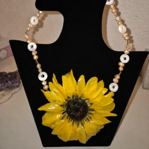 Conjunto Bahamas compuesto por una gargantilla con un gran girasol real encapsulado en resina, perlas de agua dulce y discos de nácar, junto a unos pendientes colgantes a juego exhibidos en un busto negro.