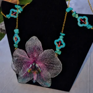Conjunto Galaxia compuesto por una gargantilla de acero dorado con gran orquídea natural fucsia y lila en resina, cuentas de turquesa y pendientes colgantes exhibido en busto negro.