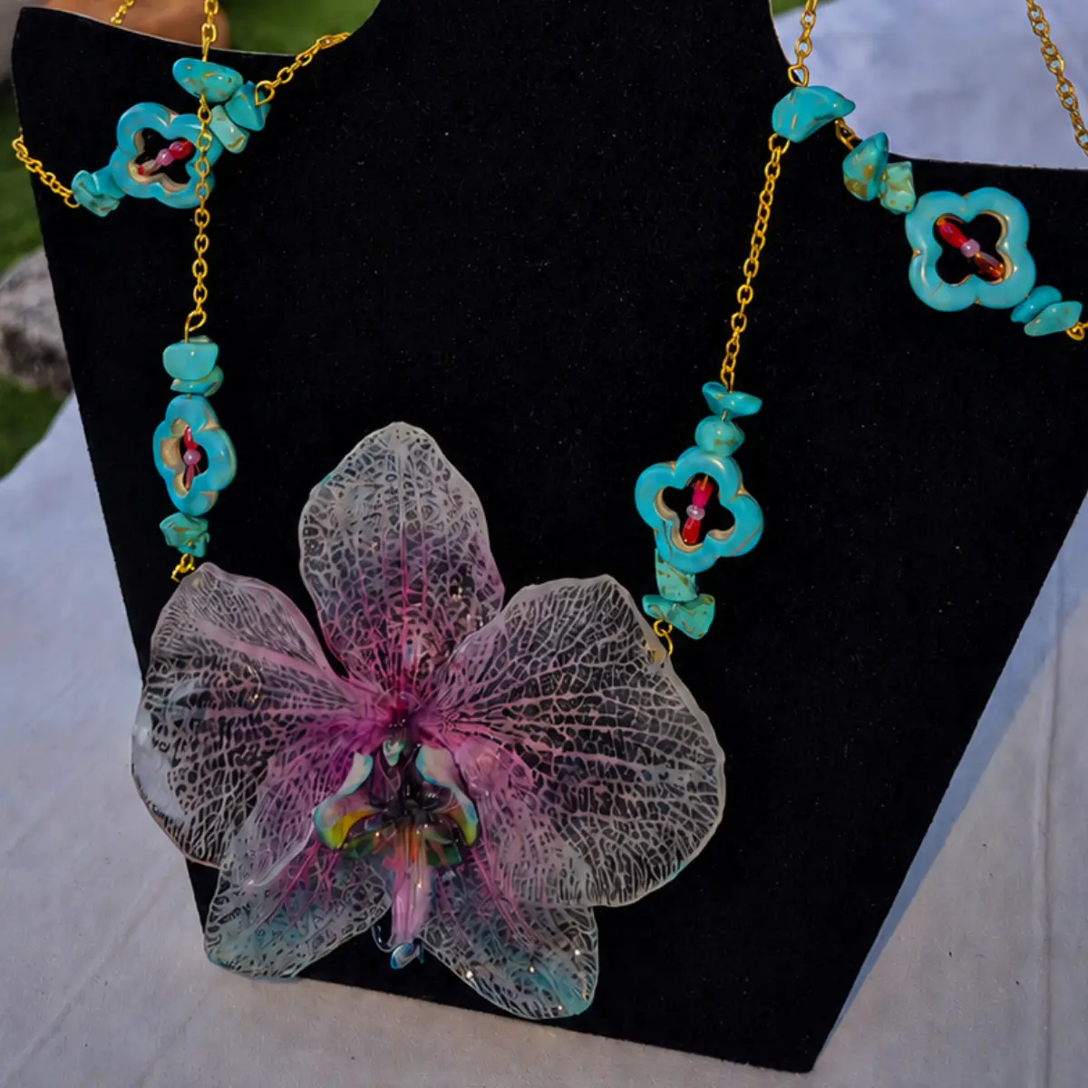 Conjunto Galaxia compuesto por una gargantilla de acero dorado con gran orquídea natural fucsia y lila en resina, cuentas de turquesa y pendientes colgantes exhibido en busto negro.
