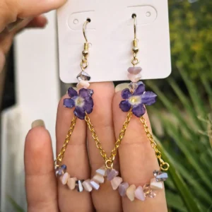 Pareja de pendientes largos de colgar con flores naturales de verbena encapsuladas en resina, unidos a una cadena dorada con cuentas de cristales irisados pastel expuestos sobre una mano.