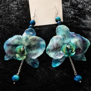 Par de pendientes colgantes artesanales de Pétalos y Glamour con una flor de orquídea natural de color azul verdoso encapsulada en resina y cuentas de turquesa, sobre fondo negro.